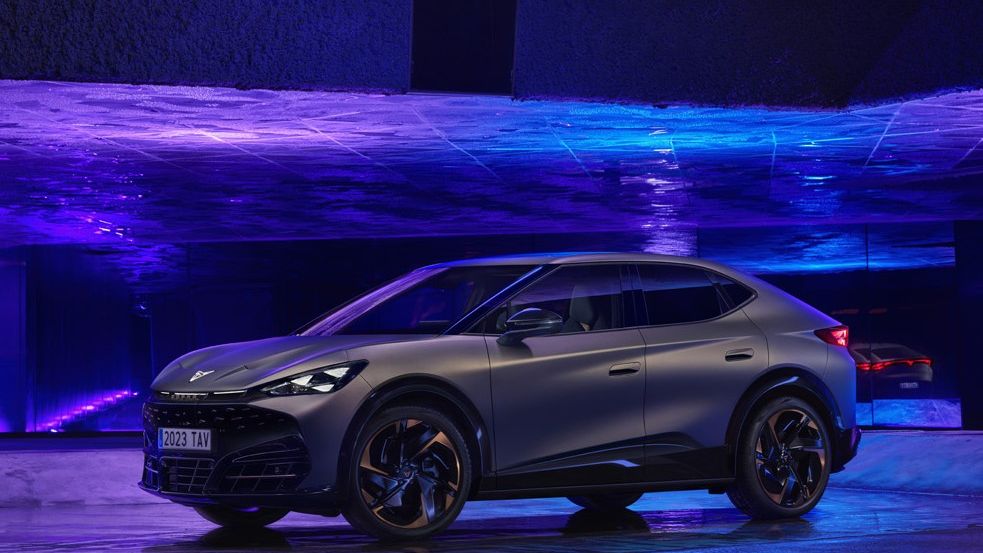 A sleek, futuristic grey CUPRA Tavascan is parked indoors against a backdrop of vibrant blue and purple lights. The stylish design features sharp lines and dark alloy wheels. Reflections of the car and lighting are visible on the polished floor.