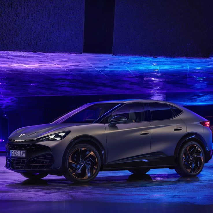 A sleek, futuristic grey CUPRA Tavascan is parked indoors against a backdrop of vibrant blue and purple lights. The stylish design features sharp lines and dark alloy wheels. Reflections of the car and lighting are visible on the polished floor.
