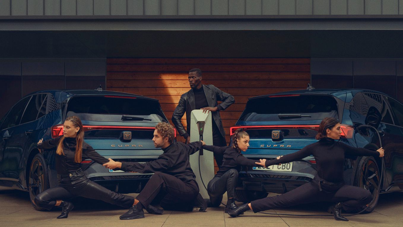 Two Aurora Blue CUPRA Born are parked side-by-side facing away, with four individuals dressed in black striking poses around them. A fifth person, also dressed in black, stands between the cars, facing the camera. All faces are blurred, maintaining their anonymity.