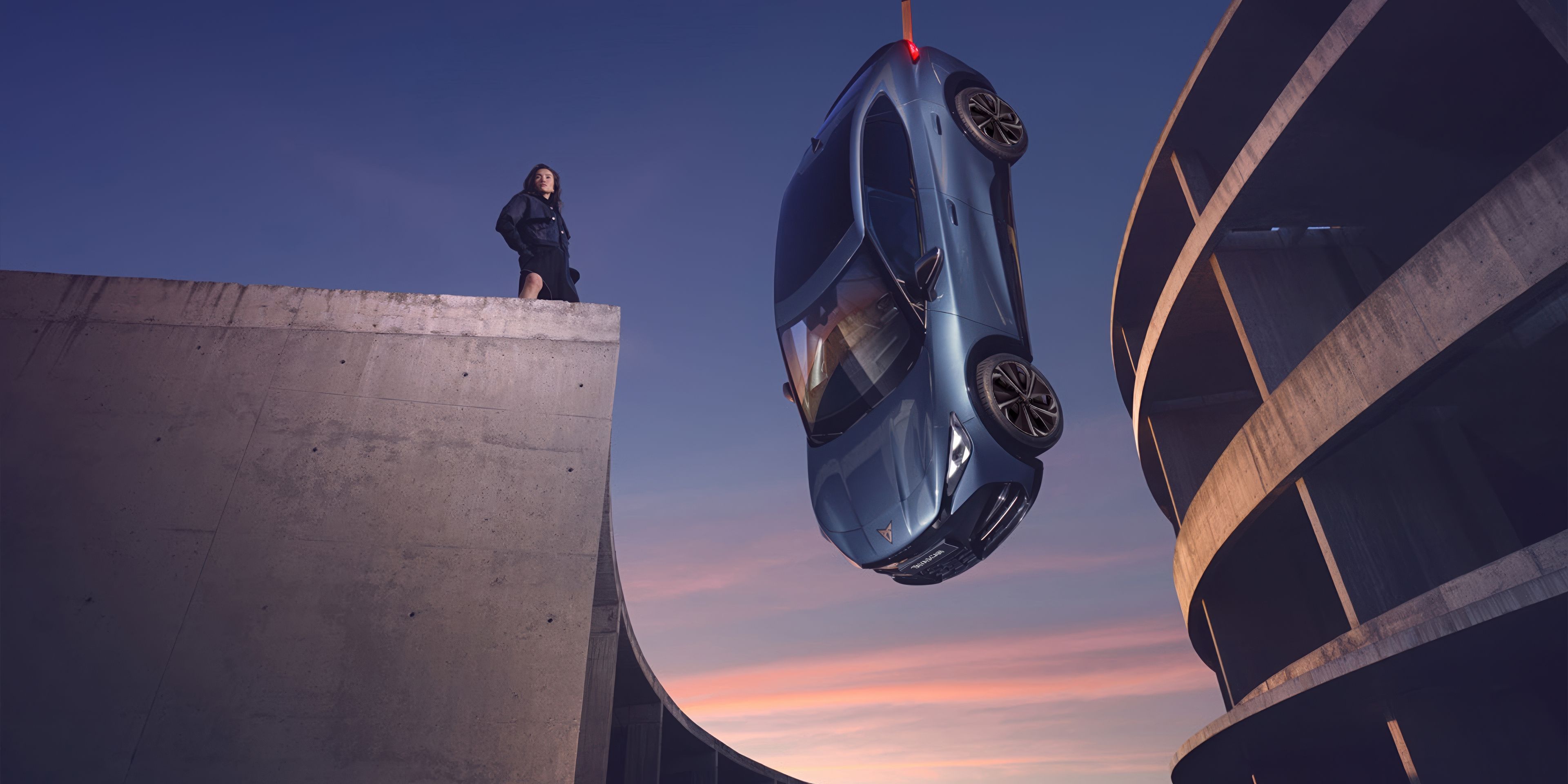 CUPRA Tavascan  is suspended by a crane over the edge of a concrete building at sunset. A lady stands on the building, observing the car as it hangs in the air. The background features parts of modern architectural structures.