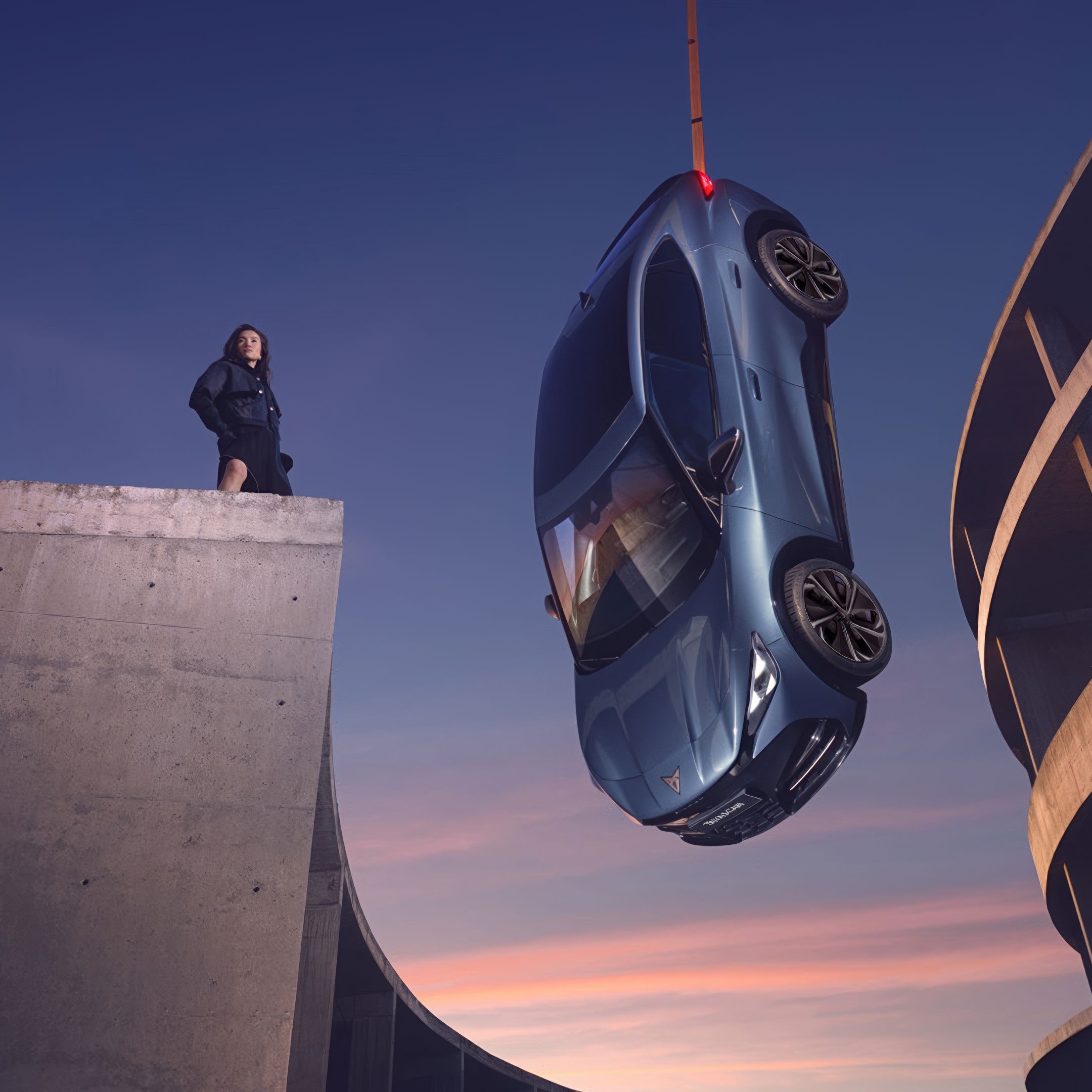 CUPRA Tavascan  is suspended by a crane over the edge of a concrete building at sunset. A lady stands on the building, observing the car as it hangs in the air. The background features parts of modern architectural structures.
