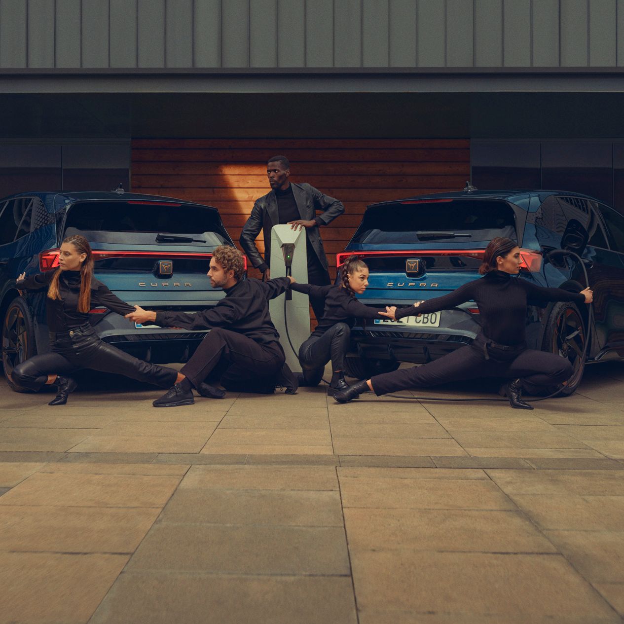 Two Aurora Blue CUPRA Born are parked side-by-side facing away, with four individuals dressed in black striking poses around them. A fifth person, also dressed in black, stands between the cars, facing the camera. All faces are blurred, maintaining their anonymity.