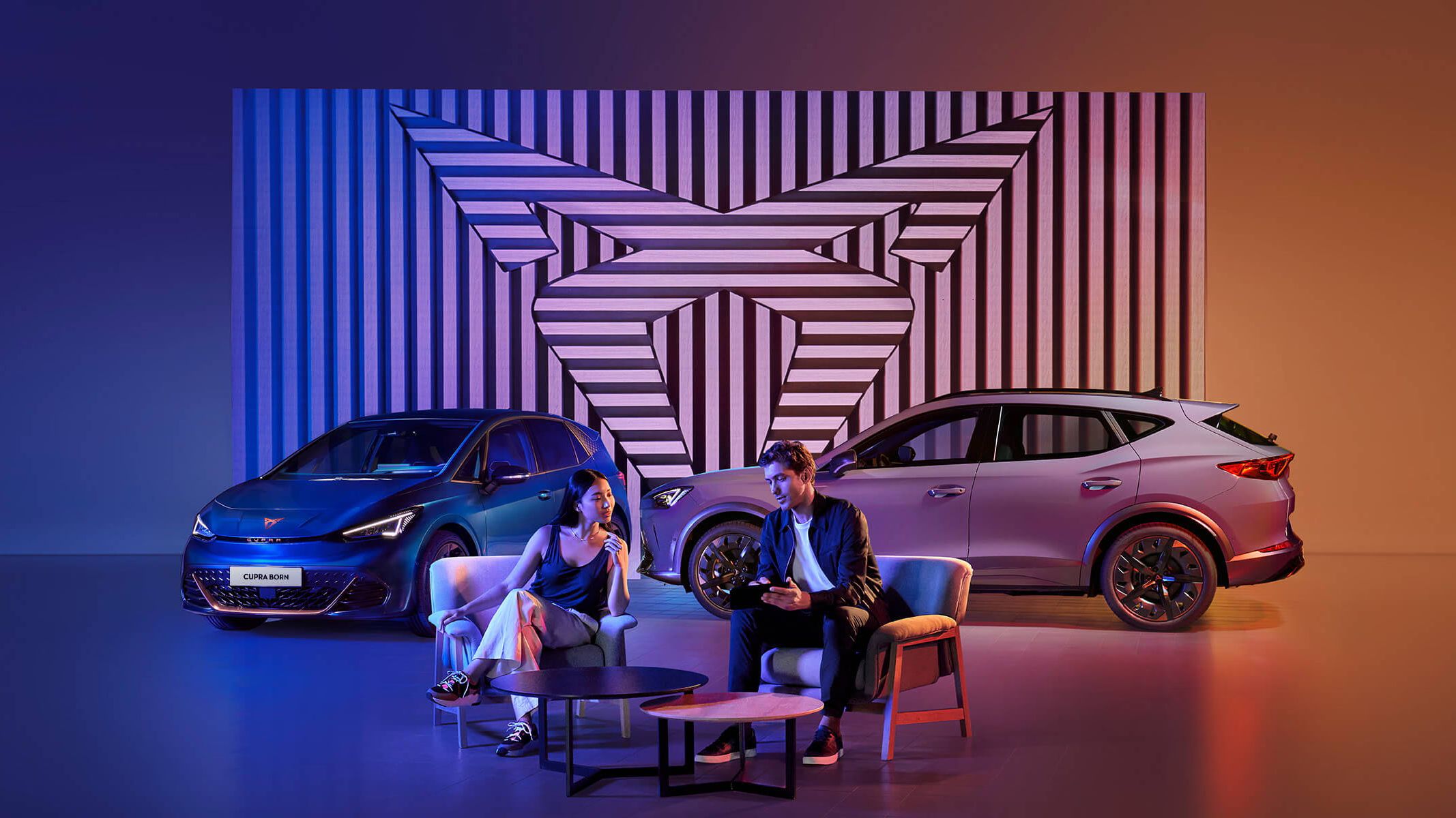 A CUPRA sales person chit chatting with a customer while sitting by the coffee table infrontr of a CUPRA Born and CUPRA Tavascan